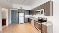 This single-floor unit presents a clean, contemporary design with a well-appointed open-concept layout connecting the kitchen and living area. The kitchen is outfitted with dark walnut-toned cabinetry, granite countertops, a white subway tile backsplash, and a full suite of stainless steel appliances including a range, over-the-range microwave, dishwasher, and refrigerator. Track lighting runs along the kitchen ceiling, providing focused task illumination throughout the space.

Light-toned hardwood flooring extends seamlessly from the kitchen through the living area, contributing to a bright, cohesive aesthetic. The living area benefits from abundant natural light through floor-to-ceiling sliding glass doors dressed with vertical blinds, with a ceiling fan overhead enhancing comfort. A secondary room, accessible via an open doorway, also features a window and ceiling fixture, offering versatility as a bedroom or flex space.

The bathroom is finished with large-format tile flooring and wall surrounds, a granite-topped vanity with dark wood cabinetry, an undermount sink, and a full soaking tub with a shower. Throughout the unit, the neutral white walls, consistent flooring, and modern fixtures combine to create a clean, move-in-ready living environment.