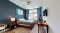 Located at 3000 Fast Ln in Fort Mill, SC, this single-floor space offers a thoughtfully designed layout that balances modern aesthetics with functional living. The bedroom serves as the focal point of the space, showcasing a striking deep teal accent wall that adds depth and character to the room. Large windows invite abundant natural light while framing views of the surrounding Fort Mill skyline, complemented by bold shibori-patterned curtains in blue and white. Light hardwood flooring runs throughout, providing a warm, cohesive foundation. The bedroom is well-appointed with a queen bed, a rustic wooden work desk, coordinating nightstands, and a ceiling fan for year-round comfort. A wall-mounted flat-screen TV and a woven accent chair add practical and decorative touches. A dedicated hallway connects the bedroom to the rest of the unit, which includes a spacious, accessibility-conscious bathroom. The bathroom features grab bars, a curtained shower with classic white subway tile, wood-look flooring, and a granite-topped vanity, prioritizing both safety and style. The overall ambiance of the space is contemporary and inviting, with carefully curated furnishings and a cohesive color palette that creates a comfortable and visually engaging environment.