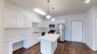 This single-floor residence offers a clean, modern aesthetic anchored by a thoughtfully designed open-concept kitchen. The kitchen is a standout feature, showcasing crisp white shaker-style cabinetry that extends to the upper walls, providing generous storage throughout. A central island with a smooth white countertop serves as a natural gathering point, offering additional workspace and casual seating potential. Stainless steel appliances — including a refrigerator, electric range, and over-the-range microwave — lend a polished, contemporary feel. A double basin sink is neatly integrated beneath the cabinetry along the perimeter wall, complemented by matching white countertops. Rich, dark hardwood-style flooring runs throughout, creating a striking contrast against the white cabinetry and light gray walls. Pendant lighting fixtures hang above the island, adding warm, focused illumination that enhances the kitchen's inviting atmosphere. The overall design emphasizes functionality and style, with a neutral color palette and quality finishes that create a bright, welcoming environment. The open layout allows for a natural flow between spaces, making this residence well-suited for both everyday living and entertaining.