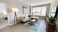 This well-appointed single-floor residence offers a thoughtfully designed layout that balances comfort and modern style. The living area is bright and welcoming, featuring large windows that frame outdoor views of a communal pool area, allowing generous natural light to fill the space. Light wood-tone flooring runs throughout the main living areas, complementing the neutral gray-beige wall palette.

The kitchen is efficiently designed with flat-front cabinetry in a warm cream finish, stainless steel appliances including a refrigerator, range, and microwave, and a white countertop peninsula that accommodates three bar stools — ideal for casual dining or entertaining.

Two bedrooms provide comfortable retreats; one features sliding glass door access to an outdoor space with pool views, while the other offers a quieter, carpeted environment with soft, warm lighting. Both rooms feature neutral tones and plush carpet flooring.

The two bathrooms are distinctly finished: one includes a full soaking tub with shower combination and tile flooring, while the other features a glass-enclosed walk-in shower, a wide floating vanity with a large mirror, and tile floors. Together, the rooms create a cohesive, contemporary living environment with clean lines and practical design throughout.