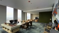 This sophisticated single-floor lounge offers an inviting and thoughtfully designed environment ideal for gathering, relaxing, or working. The space is defined by dramatic floor-to-ceiling windows that flood the interior with natural light and frame impressive panoramic city views, creating a striking visual backdrop throughout the day. The flooring features a patterned tile or carpet in muted blue-green tones, adding texture and depth to the overall aesthetic. Seating arrangements are varied and carefully curated, including plush sofas, classic leather wingback chairs, and wooden accent chairs, all arranged to encourage both intimate conversation and open social interaction. A wall-mounted flat-screen television is integrated into a deep olive-green accent wall, which serves as a bold architectural focal point and anchors one end of the room. Recessed lighting in the smooth white ceiling complements the abundant natural light, ensuring the space remains well-lit and welcoming at all hours. The overall design blends contemporary and mid-century modern influences, with a warm, earthy color palette of browns, greens, and neutrals that creates a cohesive and comfortable atmosphere throughout.