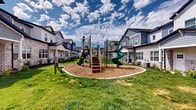 This two-story townhome community is thoughtfully designed to foster a sense of neighborhood living, combining attractive architecture with well-appointed shared outdoor spaces. The homes feature a charming mix of exterior materials, including board-and-batten siding, horizontal lap siding in neutral tones of gray and white, and stone veneer accents, giving each unit a distinctive yet cohesive curb appeal. Covered entryways with front porches invite residents to enjoy the outdoors from their own doorstep.

The community's central outdoor amenities are a standout feature. A dedicated playground area sits at the heart of the development, surfaced with wood chip mulch and equipped with a multi-level play structure featuring spiral slides, climbing stairs, and elevated platforms — ideal for younger residents. Adjacent to the playground, a communal gathering area provides green bench seating and open lawn space for relaxed outdoor enjoyment.

A separate outdoor entertaining zone includes a built-in BBQ grill station flanked by green picnic tables, creating a welcoming space for community cookouts and social gatherings. Wide concrete walkways connect all areas of the community, lined with fresh landscaping including ornamental grasses, shrubs, and mulched garden beds. The overall ambiance is bright, open, and family-friendly, reflecting a well-planned residential environment designed for active community living.