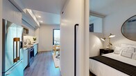 This single-floor unit showcases a thoughtfully designed open-concept layout that maximizes every inch of available space. Light wood-tone flooring flows continuously throughout, lending visual cohesion and warmth to the clean, modern aesthetic defined by crisp white walls and recessed lighting.

The kitchen is positioned along one wall and features flat-front cabinetry in a soft grey finish, stainless steel appliances including a range, microwave, and dishwasher, and under-cabinet lighting that highlights the streamlined countertops. The layout transitions naturally into a compact dining area and then into a living space anchored by a teal velvet sofa, a bold decorative rug, and a large-format artwork. Floor-to-ceiling glass doors open to a balcony, inviting abundant natural light and outdoor views of the surrounding urban streetscape.

The sleeping area is partially enclosed by a partial-height partition wall with a clerestory window above, maintaining an open feel while offering a defined, private space. A full bathroom features neutral, horizontally stacked tile surrounding a soaking tub-shower combination, light tile flooring, and recessed overhead lighting for a clean, spa-inspired finish.
