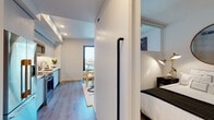 This single-floor unit showcases a thoughtfully designed open-concept layout that maximizes every inch of available space. Light wood-tone flooring flows continuously throughout, lending visual cohesion and warmth to the clean, modern aesthetic defined by crisp white walls and recessed lighting.

The kitchen is positioned along one wall and features flat-front cabinetry in a soft grey finish, stainless steel appliances including a range, microwave, and dishwasher, and under-cabinet lighting that highlights the streamlined countertops. The layout transitions naturally into a compact dining area and then into a living space anchored by a teal velvet sofa, a bold decorative rug, and a large-format artwork. Floor-to-ceiling glass doors open to a balcony, inviting abundant natural light and outdoor views of the surrounding urban streetscape.

The sleeping area is partially enclosed by a partial-height partition wall with a clerestory window above, maintaining an open feel while offering a defined, private space. A full bathroom features neutral, horizontally stacked tile surrounding a soaking tub-shower combination, light tile flooring, and recessed overhead lighting for a clean, spa-inspired finish.