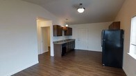 This single-story space features an open-concept layout that seamlessly connects the kitchen and living area, creating a functional and cohesive living environment. The kitchen is equipped with rich, dark wood cabinetry that extends to the upper and lower sections, providing ample storage. A granite-style countertop runs along the base cabinets, complemented by a built-in sink and a black dishwasher. A black refrigerator anchors one side of the kitchen, contributing to a consistent, coordinated appliance finish throughout. Pendant lighting fixtures hang above the kitchen area, adding focused illumination, while a ceiling-mounted light fixture serves the broader open space. The flooring throughout is a dark hardwood-style material that adds warmth and visual continuity across the entire area. The walls are finished in a clean, neutral white tone, enhancing the sense of openness. An arched doorway leads to an additional room or hallway beyond the kitchen, suggesting further living or bedroom space. The overall design is practical and modern, with a neutral palette that allows for versatile interior styling.