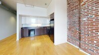 This single-floor loft-style space masterfully combines industrial heritage with contemporary design. The open layout is anchored by warm hardwood flooring that runs throughout the main areas, reflecting natural light and lending a clean, inviting atmosphere to the space.

Exposed red brick walls are a defining architectural feature, adding rich texture and a sense of history to the interior. Overhead, exposed metallic ductwork runs prominently across the ceiling, reinforcing the industrial aesthetic while contributing to the generous vertical scale of the space.

The kitchen area is well-appointed with sleek, high-gloss white upper cabinetry contrasted by rich dark lower cabinets and dark countertops. Black appliances, including a refrigerator, range, and dishwasher, complete the functional and stylish kitchen setup. The kitchen sits adjacent to the exposed brick wall, creating a compelling visual contrast between raw and refined elements.

The main open area benefits from natural light streaming through a window fitted with horizontal blinds, offering views to the outdoors. A secondary open zone with smooth white walls and the same hardwood flooring provides a versatile, neutral space.

The bathroom is compact and clean, featuring tile flooring and modern fixtures in a simple, utilitarian design. Overall, this space offers a distinctive blend of raw industrial character and polished modern finishes.