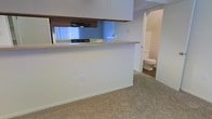 This two-floor space offers a practical and functional layout designed for everyday living. The kitchen is highlighted by a pass-through counter that opens to the adjacent living area, creating an open and connected feel between spaces. Upper cabinetry in warm wood tones provides ample storage, while an overhead range hood adds to the kitchen's utility. The light-colored countertop along the pass-through offers additional workspace or casual dining options. Natural light filters into the kitchen through vertical blinds, brightening the area with a soft, diffused glow. The flooring throughout features a speckled, neutral-toned carpet that adds a subtle texture to the space. A hallway visible from the main living area leads conveniently to a bathroom, which is outfitted with standard fixtures and maintains the clean, simple aesthetic found throughout the rest of the space. The overall color palette is light and neutral, with white walls that enhance the sense of openness. Spanning two floors, this space provides room for distinct living and private areas, making it a versatile and efficient layout suited for a variety of needs.