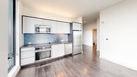 Located in Boston's East Boston neighborhood, this modern 2-bedroom, 1-bathroom residence offers a well-designed single-floor layout with contemporary finishes throughout. Hardwood flooring flows seamlessly across every room, complementing the clean, white walls and minimalist aesthetic.

The combined kitchen and living area serves as the heart of the home. The kitchen is outfitted with sleek white cabinetry, a light blue tile backsplash, stainless steel appliances including a full-size refrigerator, built-in oven, microwave, and dishwasher, along with a dark countertop that adds contrast to the bright space.

Both bedrooms are generously proportioned and bathed in natural light from expansive floor-to-ceiling windows with black frames. The primary bedroom features a corner window configuration, offering panoramic views of the Boston cityscape and harbor. The second bedroom also benefits from large windows framing striking waterfront vistas.

The bathroom is finished with large-format wood-look wall tiles in the soaking tub and shower surround, complemented by a modern floating vanity with a dark countertop and a wide mirror. A hallway connects all rooms, ensuring a practical and efficient layout throughout this well-appointed urban residence.