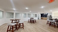 Located at 3425 Eischen Ct in Duluth, MN, this single-floor community amenity space offers a well-appointed and versatile environment designed for gathering and socializing. The layout flows between two distinct areas, each finished with light-toned luxury vinyl plank flooring that gives the space a clean, modern aesthetic throughout.

The lounge area is bright and welcoming, featuring large windows that invite abundant natural light and offer views of the surrounding exterior. Mid-century modern-inspired seating, including cushioned sofas and a round coffee table, create a comfortable and stylish setting for relaxation or casual conversation. A staircase with a sleek black metal railing connects this area to the rest of the building.

The open gathering room is spacious and versatile, lined with recessed lighting and white walls accented by a row of framed window openings that add architectural interest. High-top bistro tables with upholstered stools are arranged throughout, making the area ideal for social events or communal use.

Adjacent to the gathering room is a functional kitchenette, highlighted by a bold black hexagonal tile backsplash, natural wood cabinetry, a stainless steel refrigerator, and a wall-mounted mini-split unit. This well-designed amenity space combines practicality with contemporary style, creating an inviting hub for residents at this Duluth community.