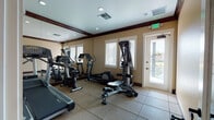 Located at 350 34th Ave Dr E in Bradenton, Florida, this single-story fitness facility offers a thoughtfully organized space designed to support a well-rounded exercise routine. The layout is divided into distinct functional zones, seamlessly connected by a transitional corridor with rich wood-toned flooring, white paneled doors, and warm neutral walls that establish a clean, welcoming atmosphere throughout.

The fitness room serves as the centerpiece of the facility, outfitted with a selection of cardio and strength-training equipment, including treadmills, elliptical machines, a multi-station cable machine, and a rowing-style trainer. Large floor-to-ceiling mirrors line one wall, visually expanding the space and allowing users to monitor their form during workouts. Durable rubberized tile flooring provides a safe, cushioned surface underfoot, while recessed lighting and crown molding with dark wood accents lend the room a polished, finished look. Multiple windows admit natural light and offer exterior views, and a glass-paneled door provides direct outdoor access.

Adjacent to the fitness room, a hallway leads to restroom facilities, complete with a wooden bench for convenience. The overall design balances functionality with a refined aesthetic, making this a practical and comfortable destination for fitness activities.