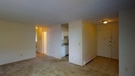 This single-floor unit offers a straightforward, functional layout with an open-concept living and dining area that connects seamlessly to the kitchen. The living space features neutral-toned carpeting and light-colored walls, creating a clean, understated backdrop. A baseboard heating unit runs along the exterior wall, indicating a practical heating setup throughout the unit.

The kitchen is partially enclosed by a dividing wall, providing a degree of separation while maintaining an open feel to the main living area. White cabinetry lines the upper portion of the kitchen, complemented by a white appliance suite including a dishwasher. A countertop with a sink is visible, offering standard kitchen functionality in a compact, efficiently arranged space.

A hallway visible from the main living area leads to additional rooms, with overhead lighting providing consistent illumination throughout. A window along the hallway wall allows some natural light to filter in. The unit's overall palette of whites and neutral tones, combined with flush-mount ceiling fixtures, gives the space a modest, clean character well-suited for everyday living.