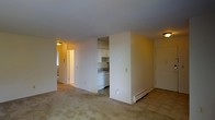 This single-floor unit offers a straightforward, functional layout with an open-concept living and dining area that connects seamlessly to the kitchen. The living space features neutral-toned carpeting and light-colored walls, creating a clean, understated backdrop. A baseboard heating unit runs along the exterior wall, indicating a practical heating setup throughout the unit.

The kitchen is partially enclosed by a dividing wall, providing a degree of separation while maintaining an open feel to the main living area. White cabinetry lines the upper portion of the kitchen, complemented by a white appliance suite including a dishwasher. A countertop with a sink is visible, offering standard kitchen functionality in a compact, efficiently arranged space.

A hallway visible from the main living area leads to additional rooms, with overhead lighting providing consistent illumination throughout. A window along the hallway wall allows some natural light to filter in. The unit's overall palette of whites and neutral tones, combined with flush-mount ceiling fixtures, gives the space a modest, clean character well-suited for everyday living.
