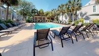 Located at 1742 Sam Rittenberg Blvd in Charleston, South Carolina, this outdoor amenity area offers a well-appointed residential pool and recreation space designed for relaxation and leisure. The centerpiece is a generously sized swimming pool with crystal-clear turquoise water, accessible via stainless steel handrails and surrounded by a broad, light-toned concrete sun deck. The deck is furnished with multiple lounge chairs and chaise lounges, providing ample space for sunbathing, as well as umbrella-shaded bistro-height tables and seating for casual outdoor dining and socializing. The area is enclosed by a sleek black iron fence with a gated entry, ensuring privacy and security. Lush, mature landscaping surrounds the perimeter, featuring tall palm trees, flowering shrubs, and manicured greenery that lend a distinctly Southern coastal character to the setting. A charming single-story clubhouse pavilion with white trim, large windows, and a pergola-style overhang anchors one end of the pool area, offering a shaded retreat. The overall ambiance is bright, open, and inviting, bathed in abundant natural sunlight with a relaxed, resort-inspired atmosphere well-suited to the warm Charleston climate.