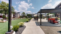 Located at Bellamy Place in Louisville, Kentucky, this thoughtfully designed outdoor community space blends modern residential living with the character of its surrounding urban environment. The grounds are anchored by expansive, well-maintained green lawns that provide open recreational space, complemented by carefully curated landscaping beds featuring flowering shrubs, ornamental grasses, and decorative stone mulch that guide visitors along smooth concrete walkways.

A central courtyard serves as the social hub of the property, outfitted with multiple outdoor dining sets shaded by large market umbrellas, sun loungers, pergola structures, and grilling stations — creating an inviting setting for gathering and relaxation. The area is framed by a striking multi-story historic brick industrial building, whose large grid windows and robust masonry provide a dramatic architectural backdrop that contrasts with the contemporary residential buildings on the opposite side.

The modern residential buildings feature a neutral exterior palette of gray and white siding with dark accent trim, private balconies, and brick detailing at the base. Mature trees are interspersed throughout the property, offering natural shade. Ample on-site parking, wide paved drives, and a gated recreational court round out the amenities, making this a comprehensive and well-connected outdoor living environment.