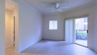 This single-floor space presents a clean, open layout designed for comfort and functionality. The main living area features light-toned ceramic tile flooring that spans the room, providing a durable and easy-to-maintain surface. Smooth, neutral-colored walls create a bright and airy atmosphere, enhanced by natural light streaming in through a window fitted with horizontal blinds. A ceiling fan is centrally mounted on the textured ceiling, offering both practical air circulation and a subtle design element. A set of vertical-blind-covered sliding glass doors opens directly to an outdoor area, seamlessly connecting the interior to the exterior and allowing for additional natural light and ventilation. The overall ambiance of the space is understated and versatile, offering a neutral canvas well-suited for a variety of uses and interior design preferences. The straightforward, uncluttered layout makes efficient use of the available space, while the combination of natural light sources and the ceiling fan contributes to a comfortable and inviting environment throughout the day.