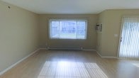 This two-floor space offers a clean, functional layout with practical features throughout. The main living area is characterized by light-toned hardwood flooring that reflects natural light, contributing to a bright and open atmosphere. A large picture window dressed with vertical blinds serves as the focal point of the room, allowing ample natural light to filter in while offering views of the surrounding greenery outdoors. Neutral beige walls provide a versatile backdrop adaptable to a variety of interior styles. Climate control is well-equipped, with a baseboard heating unit running beneath the window and a wall-mounted air conditioning unit ensuring comfortable temperatures across all seasons. Clean white baseboards frame the room neatly, adding a finished look to the space. The overall ambiance is simple and understated, offering a blank canvas ready for personalization. Spanning two floors, the space provides separation between living and private areas, making it a practical and straightforward option for everyday living.