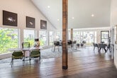 Located at 1630 Orchard Drive in Yorba Linda, California, this single-story space combines striking architectural design with modern, functional interiors. The entrance greets visitors with soaring vaulted ceilings, large-format tile flooring, a statement geometric pendant light, and a distinctive reclaimed wood structural column that anchors the space visually. Full-height glass doors invite abundant natural light and frame views of the surrounding greenery.

The expansive open-plan main area transitions seamlessly between multiple seating and gathering zones, all united by wide-plank hardwood flooring and crisp white walls. A dedicated lounge area features large picture windows overlooking a resort-style pool, filling the room with sunlight and outdoor views. A bar-height dining table anchored beneath a dramatic vaulted ceiling section offers additional flexible seating, complemented by curated artwork and decorative rugs throughout.

A compact, well-appointed kitchen includes white flat-panel cabinetry extending to the ceiling, light quartz countertops, a stainless steel sink, and stainless appliances. A clean, modern bathroom features a wide vanity with quartz counters, a large mirror, and a combination tub and shower with a window providing natural light. A private office nook is also accessible from the main entry area, completing this versatile and thoughtfully designed community space.