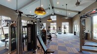 This fitness center in Humble, TX is a thoughtfully designed, single-floor facility that combines functionality with an upscale aesthetic. The space is anchored by dramatic vaulted ceilings with exposed beams, which lend a sense of openness and grandeur. Striking bronze dome pendant lights hang throughout, adding a refined, modern touch to the interior.

The main workout floor is covered in a patterned, multi-tone carpet tile that delineates zones throughout the space. Large windows along multiple walls flood the interior with natural light while offering views of the surrounding landscaped grounds. Roller shades provide adjustable light control for varying workout conditions.

The cardio zone features a row of treadmills, ellipticals, and stair climbers, each equipped with individual screens for entertainment during workouts. Cycling bikes and additional cardio equipment occupy a central area, while a dedicated strength training section houses a multi-station cable machine, weight benches, and free weights. A punching bag station and wall-mounted pull-up/dip rack provide a combat and functional fitness option. Wall-mounted televisions are positioned throughout the space for added convenience.

The entry area includes a hand sanitizer station, posted fitness rules, and a large mirror that enhances the sense of depth. The overall ambiance is energetic yet polished, making this fitness center a premium amenity.
