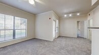 This single-floor residential unit is thoughtfully laid out with a clean, neutral aesthetic carried throughout. The main living area is generously sized, featuring plush wall-to-wall carpeting, crown molding, and a ceiling fan overhead. Multiple windows fitted with blinds allow natural light to filter in, offering views of surrounding greenery. A sliding glass door and a separate entry door provide convenient access points. Track lighting near the entryway adds a functional accent.

The kitchen is equipped with white cabinetry, a dark laminate countertop, and a full suite of black appliances including an electric range, over-the-range microwave, and refrigerator. Light wood-look flooring runs through the kitchen, which opens to the main living space via a breakfast bar-style counter. A dedicated bedroom offers carpeted flooring, a ceiling fan, and a window for natural light.

The bathroom presents a wide vanity with warm-toned countertops, white cabinetry, a large mirror with overhead lighting, and a tiled shower-tub combination. Light wood-look flooring ties the space together. Additionally, the unit includes an in-unit laundry area with a washer and wall-mounted shelving, adding everyday convenience to this well-appointed, move-in-ready space.