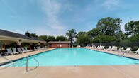 This outdoor recreational space centers around a generously sized swimming pool with clear, blue-green water and a classic rectangular design. The pool deck is finished in a warm concrete surround, offering plenty of open space for movement and relaxation. Numerous white lounge chairs are arranged along multiple sides of the pool, providing abundant seating for sunbathing and leisure. A brick pool house structure serves as the functional hub of the area, housing what appears to be restrooms and utility facilities, and is accented by a mounted life preserver ring for safety. To one side, a charming covered pavilion with a pitched roof offers a shaded retreat from the sun. The entire pool area is enclosed by a metal perimeter fence, ensuring privacy and security, with access controlled through a dedicated pool gate. Lush, mature trees frame the background on all sides, creating a natural canopy that enhances the sense of seclusion and greenery. The space benefits from abundant natural light under open skies, making it an ideal setting for outdoor recreation. A posted sign at the entrance reminds visitors to use the designated pool gate, reflecting the well-managed nature of this community amenity.