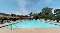 This outdoor recreational space centers around a generously sized swimming pool with clear, blue-green water and a classic rectangular design. The pool deck is finished in a warm concrete surround, offering plenty of open space for movement and relaxation. Numerous white lounge chairs are arranged along multiple sides of the pool, providing abundant seating for sunbathing and leisure. A brick pool house structure serves as the functional hub of the area, housing what appears to be restrooms and utility facilities, and is accented by a mounted life preserver ring for safety. To one side, a charming covered pavilion with a pitched roof offers a shaded retreat from the sun. The entire pool area is enclosed by a metal perimeter fence, ensuring privacy and security, with access controlled through a dedicated pool gate. Lush, mature trees frame the background on all sides, creating a natural canopy that enhances the sense of seclusion and greenery. The space benefits from abundant natural light under open skies, making it an ideal setting for outdoor recreation. A posted sign at the entrance reminds visitors to use the designated pool gate, reflecting the well-managed nature of this community amenity.