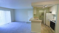 This single-floor residence offers a clean, open-concept layout that seamlessly connects the living area and kitchen. The living space is finished with plush wall-to-wall carpeting in a soft blue-grey tone, creating a comfortable and inviting atmosphere. Smooth, light-toned walls throughout the unit contribute to a bright and airy feel, complemented by a textured ceiling that adds subtle visual interest.

The kitchen is defined by a peninsula countertop with an integrated sink, providing a natural division between the cooking and living spaces while maintaining an open sightline. Upper cabinetry in a light finish offers practical storage, and track lighting above the kitchen area ensures focused illumination for meal preparation. The kitchen floor transitions to tile, offering a durable and easy-to-clean surface. A full-size refrigerator anchors the kitchen, and additional appliances are visible in a dedicated utility or laundry area adjacent to the kitchen.

A paneled interior door leads from the living area, and a floor vent indicates a forced-air heating and cooling system throughout the unit. The overall design is functional and straightforward, with a neutral color palette that makes the space adaptable to a variety of interior styles.