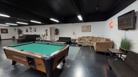 This expansive rec room offers a versatile, open-plan layout designed for entertainment and socializing. The space is defined by its striking contrast of black-painted ceilings and exposed infrastructure against bright white walls, creating a bold, industrial-modern aesthetic. Dark, polished concrete floors run throughout, lending a sleek and durable foundation that ties the entire space together.

The room is generously illuminated by rows of overhead fluorescent strip lighting, ensuring the entire area is well-lit despite the dark ceiling. A centerpiece pool table anchors one section of the space, accompanied by a cue rack for easy access. Multiple seating arrangements are distributed throughout, including a large sectional sofa, a dark leather sectional, additional sofas, and a pair of accent armchairs — providing ample space for guests to gather and relax.

A dedicated lounge nook features a wall-mounted flat-screen television alongside a neon "Superstar" sign, adding a playful, vibrant touch to the ambiance. A bar-height table with stools offers a casual spot for conversation. Decorative wall art, potted plants, and ornamental grasses are placed throughout, softening the industrial feel. A separate windowed area provides access to an outdoor deck, bringing in natural light and offering a refreshing connection to the exterior.