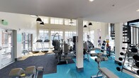 This fitness center offers a bright and well-organized workout environment designed to accommodate a variety of exercise routines. Large windows along multiple walls flood the space with abundant natural light, creating an open and energizing atmosphere. Elevated transom windows further enhance the sense of height and airiness throughout the room.

The facility is divided into distinct workout zones, each with its own flooring to delineate function. A dark rubberized mat area houses a selection of cardio machines, including treadmills and ellipticals, while a vibrant teal flooring section supports strength and free-weight training. A dedicated weight rack displays an organized array of dumbbells and medicine balls, making equipment easily accessible.

Structural columns punctuate the space, lending an architectural character without impeding the flow of the room. Overhead, ceiling fans and pendant-style black light fixtures provide both comfort and focused illumination. The clean white walls and neutral ceiling contribute to a fresh, modern aesthetic that keeps the environment feeling spacious and motivating for users at all fitness levels.