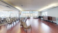 Located at 170 Erb St W in Waterloo, Ontario, this generously proportioned single-floor community space is designed to accommodate a variety of functions with comfort and elegance. The facility is divided into several distinct yet interconnected areas that together create a warm and welcoming environment.

The formal dining room is a standout feature, offering an abundance of natural light through expansive floor-to-ceiling windows that frame views of mature surrounding trees. Elegantly set round tables with white linens are illuminated by decorative pendant chandeliers, while cove lighting along the ceiling adds a refined ambiance. Neutral carpeting and classic wainscoting lend a polished, residential feel.

Adjacent to the dining area is a large activity and event hall finished with rich hardwood-style flooring. This versatile space accommodates seating arrangements for gatherings or performances and features a grand piano, large windows, and draped curtains. Cove lighting continues throughout, maintaining a consistent and inviting atmosphere.

A functional servery area supports dining operations, featuring a long service counter, built-in cabinetry, and organized storage. A casual lounge and dining nook, also with hardwood flooring, provides a more relaxed setting with informal seating and additional storage. Throughout, the design balances practicality with a warm, community-oriented aesthetic suited to daily programming and social activities.