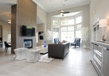 This single-floor space makes an immediate impression with its dramatic ceiling height and abundant natural light. Floor-to-ceiling windows with sleek roller shades line the main seating area, flooding the interior with daylight and offering open views of the surrounding greenery. A contemporary ceiling fan anchors the upper expanse of the room, adding both function and visual interest to the lofty overhead space.

The lounge area is thoughtfully furnished with a dark leather sofa accented by blue and white throw pillows, paired with striped upholstered accent chairs that add a refined touch. A glossy white coffee table serves as the focal centerpiece of the seating arrangement, adorned with tasteful décor including a tray and a sculptural blue glass accent piece. A soft area rug grounds the conversation area, while decorative white branch arrangements in planters add an organic, elegant contrast to the otherwise modern palette.

The neutral color scheme — featuring soft grays, whites, and warm beige tones — lends the space a clean, polished aesthetic. A frosted glass partition wall to one side subtly divides areas while maintaining an open, airy feel. Light-toned flooring and recessed lighting complete the sophisticated, welcoming ambiance of this versatile space.