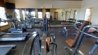 This fitness center offers a comprehensive workout environment designed to accommodate a variety of exercise routines. The space is outfitted with an extensive selection of cardio machines, including treadmills and ellipticals, neatly arranged along one side of the room to maximize floor space. Strength training equipment is well-represented, with multi-functional cable and weight machines positioned throughout the center of the room, complemented by a dedicated free weights station featuring a full dumbbell rack. Large floor-to-ceiling mirrors line an entire wall, creating a sense of expanded space while allowing users to monitor their form during workouts. The flooring consists of durable, dark rubber matting suited for high-traffic fitness use. Natural light filters in through multiple windows fitted with decorative valances, lending a bright and welcoming atmosphere to the room. The neutral-toned walls and warm accents contribute to a clean, functional aesthetic. The single-floor layout ensures easy accessibility throughout, and the open floor plan provides ample room to move freely between equipment stations, making this fitness center a practical and well-appointed facility for a full-body workout regimen.