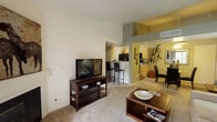 This single-floor residence offers a well-designed layout that seamlessly connects the main living areas. The living room is immediately welcoming, characterized by impressively high ceilings that lend a sense of openness and volume to the space. Soft carpeting underfoot adds warmth and comfort, while the open-concept design allows for a natural flow between the living, dining, and kitchen areas. The kitchen is equipped with warm cabinetry and features a raised breakfast bar with bar seating, making it an inviting spot for casual dining or entertaining. Adjacent to the kitchen, a dedicated dining area provides additional space for more formal meals. Throughout the interior, a neutral color palette creates a calm and versatile ambiance that complements a variety of décor styles. One of the standout features of this residence is its private balcony, accessible from the living area and framed by an elegant arched overhang. The balcony overlooks a beautifully landscaped community courtyard, complete with mature trees, manicured greenery, walking paths, and a sparkling outdoor pool — offering a serene and picturesque outdoor retreat. The combination of thoughtful interior design and appealing outdoor views makes this a comfortable and visually engaging living space.