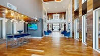 Located at 1400 Encore Place in Sanford, Florida, this single-story clubhouse is a thoughtfully designed communal facility that seamlessly blends contemporary aesthetics with inviting warmth. The space makes an immediate impression through its dramatic double-height ceilings and expansive floor-to-ceiling windows, flooding the interior with abundant natural light and offering views of the outdoor pool area beyond.

The grand entry lobby showcases rich hardwood flooring arranged in a distinctive pattern, flanked by warm wood-paneled accent walls and complemented by textured wallcoverings in cool grey tones. Bold blue upholstered seating anchors the main lounge area, while decorative wall sconces and pendant lighting add sophisticated illumination throughout.

A spacious bar-style gathering counter with a crisp white quartz surface and dark wood barstools serves as a social hub, backed by a striking three-dimensional hexagonal tile wall bathed in warm under-lighting. An intimate conference room features a round dark wood table, velvet blue chairs, a sculptural globe chandelier, and a built-in water wall feature framed in rich wood paneling.

Additional seating nooks with modern blue accent chairs and curated artwork create comfortable, private retreats throughout. The space culminates in a rear glass entry that opens directly to the pool courtyard, reinforcing a seamless indoor-outdoor connection.
