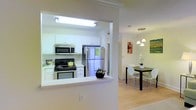 Located at 100 Adelaide Cir in Morrisville, NC, this single-story residence offers a thoughtfully designed layout that emphasizes open, connected living spaces. The kitchen is a functional centerpiece, outfitted with stainless steel appliances including a refrigerator, range with oven, and over-the-range microwave. White cabinetry with raised-panel doors lines the walls, complemented by clean white countertops that extend to a pass-through peninsula, creating a seamless connection to the adjacent dining area. The flooring throughout transitions smoothly from the kitchen into the dining space, featuring light hardwood that adds warmth and continuity. Crown molding details frame the kitchen opening, lending an elegant architectural touch. The dining area is bright and welcoming, illuminated by a set of pendant lights that hang above the dining space, creating a focused and inviting atmosphere for meals. The overall color palette is light and neutral, with white walls and trim that enhance the sense of openness. The interior maintains a clean, modern aesthetic throughout, making it both practical and visually appealing for everyday living.