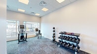 This fitness center on Uptown Ave in Broomfield, Colorado offers a clean, modern workout environment designed for functional and versatile exercise. The space is laid out on a single floor with a well-organized open floor plan that accommodates a variety of fitness activities.

The room features a drop-panel ceiling with recessed LED lighting panels that provide consistent, bright illumination throughout. Durable, multi-tone carpet tiles cover the floor, offering both comfort and traction during workouts. The walls are finished in a crisp white, contributing to the space's bright and airy feel.

Large windows along one wall allow abundant natural light to flow into the space and offer views of the surrounding Broomfield neighborhood. Cardio equipment, including an elliptical trainer and a stationary recumbent bike, is positioned near the windows to take advantage of the natural light and outdoor views.

On the opposite side of the room, a three-tier dumbbell rack holds a comprehensive set of free weights, accompanied by a medicine ball rack stocked with multiple weighted balls and resistance bands. An adjustable weight bench provides additional versatility for strength training exercises. The space also includes room for open floor movement and stretching. A secure entry door with a window panel provides convenient access to the facility.