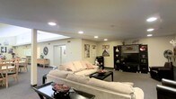 This single-floor space offers an open and functional layout designed for comfortable everyday living. The main living area is generously sized, centered around a large, plush sectional sofa in neutral tones, paired with a dark-framed coffee table that anchors the seating arrangement. Recessed lighting is installed throughout the ceiling, providing consistent and even illumination across the space. A skylight feature near the entryway adds a welcome touch of natural light, brightening the otherwise interior-facing area.

The entertainment zone is well-appointed, featuring a large flat-screen television mounted within a dark media console unit, flanked by shelving for decorative items and media storage. Movie posters adorn the walls, lending a casual, personalized character to the space. The flooring is carpeted throughout, contributing to a warm and inviting ambiance.

A hallway visible in the background leads to additional areas of the space, suggesting a thoughtful floor plan with clear circulation. Subtle decorative touches — including a starburst wall mirror, indoor greenery, and accent pieces — round out the interior. Overall, the space presents a practical and welcoming environment suited for relaxation and entertainment.