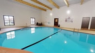 This single-floor facility offers a diverse range of amenities designed to support active wellness pursuits. The centerpiece is a generously sized indoor swimming pool, characterized by its striking turquoise water and clean, curved edges. The pool area features an elevated ceiling supported by prominent exposed wood beams, lending a warm, structural aesthetic to the otherwise crisp white-tiled walls. Entry steps with metal handrails provide safe pool access, while a modest seating area with chairs and a small table is positioned nearby, offering a practical rest space. Natural light filters in through a window along one wall, brightening the environment. Posted signage with pool rules and guidelines is clearly visible near the door.

A separate area of the facility showcases a bold, graphic mural painted directly onto concrete block walls. The design incorporates large geometric shapes in primary colors — yellow, blue, and red — set against a white background, creating a dynamic and energetic visual statement. Directional track lighting mounted overhead highlights the mural and illuminates the open floor space below. Together, the pool and activity spaces create a well-rounded, functional facility suited for both aquatic and fitness-focused activities.