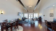 This single-story clubhouse offers an open, airy interior defined by striking architectural details and a well-appointed layout suited for community gatherings and relaxed socializing. The most prominent feature is the dramatically high vaulted ceiling, accented by bold dark exposed beams that draw the eye upward and lend an elegant yet casual character to the space. A decorative chandelier hangs centrally, complementing warm wall sconces that add a soft, inviting glow throughout.

Rich, dark hardwood flooring runs across the entire main area, grounding the open layout and providing a refined contrast to the light-toned walls. Large windows and French doors along the far wall flood the interior with natural light while offering views to the exterior, enhancing the sense of openness.

The main living area is furnished with multiple seating arrangements, including plush sofas and accent chairs anchored by area rugs, creating comfortable zones for conversation. A wall-mounted television adds a functional entertainment element. Near one side of the room, a dining table with chairs provides space for casual meals or small group activities, while a workstation area with a desktop computer offers a practical resource for residents.