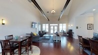 This single-story clubhouse offers an open, airy interior defined by striking architectural details and a well-appointed layout suited for community gatherings and relaxed socializing. The most prominent feature is the dramatically high vaulted ceiling, accented by bold dark exposed beams that draw the eye upward and lend an elegant yet casual character to the space. A decorative chandelier hangs centrally, complementing warm wall sconces that add a soft, inviting glow throughout.

Rich, dark hardwood flooring runs across the entire main area, grounding the open layout and providing a refined contrast to the light-toned walls. Large windows and French doors along the far wall flood the interior with natural light while offering views to the exterior, enhancing the sense of openness.

The main living area is furnished with multiple seating arrangements, including plush sofas and accent chairs anchored by area rugs, creating comfortable zones for conversation. A wall-mounted television adds a functional entertainment element. Near one side of the room, a dining table with chairs provides space for casual meals or small group activities, while a workstation area with a desktop computer offers a practical resource for residents.