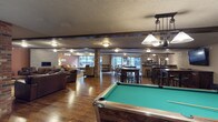 This expansive single-floor communal space is designed for recreation and socializing, combining multiple functional areas within one open-concept layout. Rich hardwood flooring runs throughout, unifying the space and adding warmth to the industrial-inspired aesthetic. Exposed beam accents and textured ceilings contribute to the room's character, while brick column details add a rugged, urban touch.

At the forefront sits a full-sized pool table, illuminated by a distinctive pulley-style pendant light fixture that serves as a focal point for the recreation area. Beyond it, a generous dining zone features dark wood tables and cross-back chairs, comfortably accommodating large groups. Large windows line the far wall, allowing natural light to fill the interior and providing views of the surrounding area.

To one side, a cozy lounge features plush sofas and armchairs arranged around a fireplace, ideal for relaxed conversation. On the opposite end, a bar counter with tall stools leads into a well-equipped kitchen area with stainless steel appliances and pendant lighting overhead. The overall ambiance is warm, social, and thoughtfully designed for communal use.
