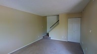 This two-story space features a spacious main living area finished with wall-to-wall carpeting in a neutral gray tone, providing a practical and comfortable foundation throughout. The walls are painted in a warm beige, creating a clean and versatile backdrop suitable for a variety of interior styles. A textured popcorn ceiling adds a classic architectural detail overhead.

A centrally positioned staircase with a dark handrail connects the main level to the upper floor, serving as a functional focal point within the living area. The staircase design is straightforward, with carpeted treads that complement the flooring on the main level. Adjacent to the staircase, a standard white paneled door provides access to an additional room or closet, contributing to the practical layout of the space.

White baseboards run along the perimeter of the room, offering a crisp contrast to the beige walls and gray carpet. Electrical outlets are evenly distributed along the walls, ensuring convenient access throughout. The overall design is clean and understated, offering a neutral, move-in-ready environment well-suited for customization.