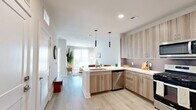 This thoughtfully designed one-bedroom, one-bathroom unit offers a bright, contemporary living experience across a single floor. The open-concept layout seamlessly connects the kitchen and living room, creating a spacious and functional common area bathed in natural light from large sliding glass doors. The kitchen is a standout feature, showcasing flat-panel wood-tone cabinetry, a quartz peninsula with bar seating, stainless steel appliances including a gas range and French door refrigerator, and sleek pendant lighting overhead. Light wood-look flooring runs throughout the unit, enhancing the clean, modern aesthetic. The living area offers a comfortable retreat with ample room for lounging and entertaining, complemented by wall-mounted media display and views to the outdoors through full-length vertical blinds. The bedroom is a calm, light-filled space with a large window, ceiling fan, and generous natural light. Three closets, including a spacious walk-in with dual hanging rods and shelving, provide exceptional storage. The bathroom features an extended dual-sink vanity with wood-tone cabinetry, a large mirror, and warm neutral finishes that echo the refined palette found throughout the home.