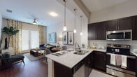 This single-floor residence offers a thoughtfully designed open-concept layout that seamlessly connects the kitchen, dining, and living areas. The kitchen is a standout feature, equipped with dark espresso cabinetry, granite countertops, stainless steel appliances — including a refrigerator, range, and built-in microwave — and a peninsula island with a double-basin sink and pendant lighting above. Rich hardwood-style flooring flows throughout the main living area, adding warmth and continuity to the space. The living room is bright and welcoming, with a ceiling fan, large windows dressed in patterned curtains, and ample room for comfortable seating. The bedroom is a calm retreat, featuring plush carpeting, generous natural light from oversized windows with blinds and sheer drapes, and a ceiling fan for year-round comfort. The full bathroom continues the design language of the home with matching dark cabinetry, a granite vanity top, tile flooring, and a full tub-shower combination. Throughout the residence, neutral walls provide a versatile backdrop, while consistent finishes in the cabinetry and hardware tie each space together into a cohesive, modern aesthetic.