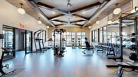 Located at 2116 Pleasant Road in Fort Mill, SC, this fitness center offers a spacious, single-floor exercise facility designed with both function and aesthetic appeal in mind. The room's most striking architectural feature is its dramatically high ceiling adorned with rich, dark wood exposed beams arranged in a coffered pattern, lending a warm, upscale character to the space. Large globe pendant lights and ceiling fans are suspended from the ceiling structure, providing both ample illumination and airflow throughout the room.

The exercise floor is finished with smooth, light-toned wood-look flooring that complements the neutral cream walls and creates a clean, modern atmosphere. Generous windows with dark-trimmed frames line the walls on multiple sides, inviting abundant natural light and offering views of the surrounding outdoor environment.

The facility is thoroughly equipped to support a wide range of fitness activities. Cardio equipment includes treadmills, stair climbers, and stationary bikes positioned to take advantage of the natural light and views. Strength training is well-represented with a cable machine, power rack with a rowing machine attachment, a full dumbbell rack, and adjustable benches. A dedicated zone near the entrance features a pull-up and dip station. Glass-paneled double doors connect sections of the facility, contributing to an open, airy feel throughout this well-appointed fitness space.