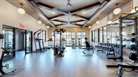 Located at 2116 Pleasant Road in Fort Mill, SC, this fitness center offers a spacious, single-floor exercise facility designed with both function and aesthetic appeal in mind. The room's most striking architectural feature is its dramatically high ceiling adorned with rich, dark wood exposed beams arranged in a coffered pattern, lending a warm, upscale character to the space. Large globe pendant lights and ceiling fans are suspended from the ceiling structure, providing both ample illumination and airflow throughout the room.

The exercise floor is finished with smooth, light-toned wood-look flooring that complements the neutral cream walls and creates a clean, modern atmosphere. Generous windows with dark-trimmed frames line the walls on multiple sides, inviting abundant natural light and offering views of the surrounding outdoor environment.

The facility is thoroughly equipped to support a wide range of fitness activities. Cardio equipment includes treadmills, stair climbers, and stationary bikes positioned to take advantage of the natural light and views. Strength training is well-represented with a cable machine, power rack with a rowing machine attachment, a full dumbbell rack, and adjustable benches. A dedicated zone near the entrance features a pull-up and dip station. Glass-paneled double doors connect sections of the facility, contributing to an open, airy feel throughout this well-appointed fitness space.