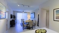 This single-floor space offers a thoughtfully designed open-concept layout that seamlessly blends living and dining areas into one cohesive environment. The dining area serves as a central focal point, featuring a round table with upholstered chairs set atop a patterned area rug, creating an inviting atmosphere for gatherings. Modern track lighting runs along the ceiling, providing adjustable illumination that complements the warm glow of accent lamps placed throughout the space. A ceiling fan adds both function and style overhead. Large sliding glass doors dressed with vertical blinds span one wall, flooding the interior with natural light and offering direct access to an outdoor area. The light-toned tile flooring extends throughout, enhancing the sense of openness and reflecting light to brighten the entire space. Neutral white walls provide a clean, versatile backdrop, while framed wall art adds character. A glimpse of the living area in the background reveals a comfortable seating arrangement, and lush greenery in the corner introduces a touch of nature indoors. The overall ambiance is bright, airy, and welcoming, with a modern yet comfortable aesthetic throughout.