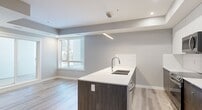 This West Garneau unit offers a thoughtfully designed, single-floor layout accommodating two bedrooms, one bathroom, and a combined kitchen and living area. Light-toned wood-style flooring runs throughout, creating visual continuity across the space.

The kitchen is a standout feature, showcasing a large island with a dark wood-paneled base and white quartz-style countertop, an undermount sink, and stainless steel appliances including a refrigerator, range, and built-in microwave. Upper cabinetry combines white flat-panel doors with dark wood lower cabinets, and recessed lighting with a tray ceiling detail adds a refined touch.

The primary bedroom benefits from natural light via a large window and sliding glass door, providing outdoor access. It includes a built-in closet with shelving. The secondary bedroom is a compact, neutral-toned room with a sliding closet.

The hallway connects the rooms efficiently with clean, gray-painted walls and white trim. The bathroom features large-format white tile flooring, a soaker tub with shower, a dark wood vanity with a white countertop, and recessed lighting, maintaining the unit's consistent modern aesthetic.