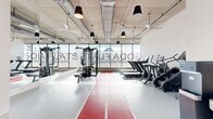 Located at 2820 W Broad St in Richmond, VA, this fitness center at Soda Flats offers a well-appointed exercise room designed for a comprehensive workout experience. The space features an industrial-modern aesthetic, highlighted by an exposed concrete ceiling with visible ductwork, pendant lighting, and ceiling fans that maintain airflow throughout. A bold red indoor running track with white lane markings runs the length of the floor, serving as both a functional and striking design element against the light gray flooring. Floor-to-ceiling windows along one wall flood the space with natural light and provide open sky views, creating an energizing atmosphere. Full-length mirrors are positioned along a side wall to maximize the sense of space and support proper form during exercise. The room is equipped with a multi-station cable and strength training machine, a rack of dumbbells with an accompanying bench, and a row of cardio machines including treadmills and ellipticals. A water fountain and water bottle filling station are conveniently located near the entrance. The clean white walls and bright, airy environment contribute to a motivating and modern fitness atmosphere suited for a variety of workout routines.