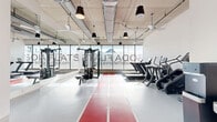Located at 2820 W Broad St in Richmond, VA, this fitness center at Soda Flats offers a well-appointed exercise room designed for a comprehensive workout experience. The space features an industrial-modern aesthetic, highlighted by an exposed concrete ceiling with visible ductwork, pendant lighting, and ceiling fans that maintain airflow throughout. A bold red indoor running track with white lane markings runs the length of the floor, serving as both a functional and striking design element against the light gray flooring. Floor-to-ceiling windows along one wall flood the space with natural light and provide open sky views, creating an energizing atmosphere. Full-length mirrors are positioned along a side wall to maximize the sense of space and support proper form during exercise. The room is equipped with a multi-station cable and strength training machine, a rack of dumbbells with an accompanying bench, and a row of cardio machines including treadmills and ellipticals. A water fountain and water bottle filling station are conveniently located near the entrance. The clean white walls and bright, airy environment contribute to a motivating and modern fitness atmosphere suited for a variety of workout routines.
