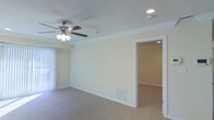 This single-floor residence offers a clean, well-maintained interior with a comfortable and functional layout. The main living area is spacious and inviting, featuring soft wall-to-wall carpeting that adds warmth underfoot. Crown molding runs along the ceiling perimeter, lending a refined, classic touch to the space. A ceiling fan with an integrated light fixture provides both air circulation and ambient lighting, while recessed lighting adds additional illumination throughout.

Natural light filters into the living area through a large window dressed with vertical blinds, creating a bright and airy atmosphere. A floor-level HVAC vent ensures efficient climate control throughout the space. The neutral, light-toned walls provide a versatile backdrop well-suited for a variety of interior styles.

An open doorway connects the main living area to an adjacent room, also carpeted and lit with recessed lighting, suggesting a seamless flow between living spaces. The overall design is understated and practical, with consistent finishes throughout that contribute to a cohesive and welcoming environment. This single-level layout offers ease of accessibility and a straightforward, comfortable living experience.